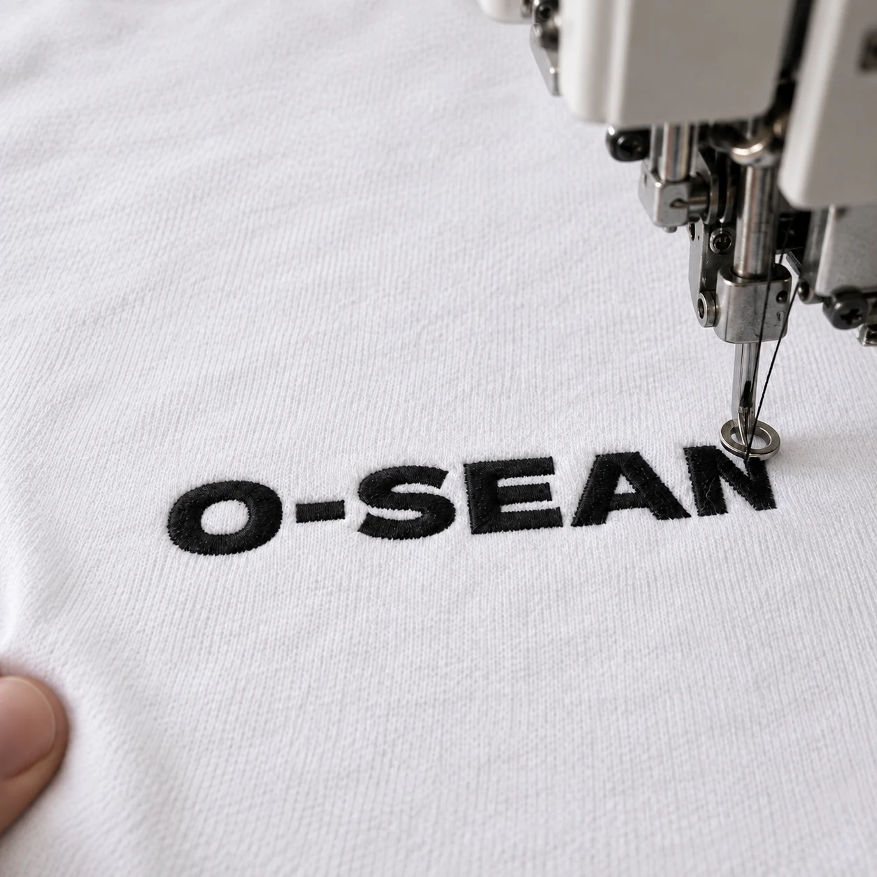 Embroidery process: a machine stitching the O-SEAN logo in black thread on a white T-shirt
