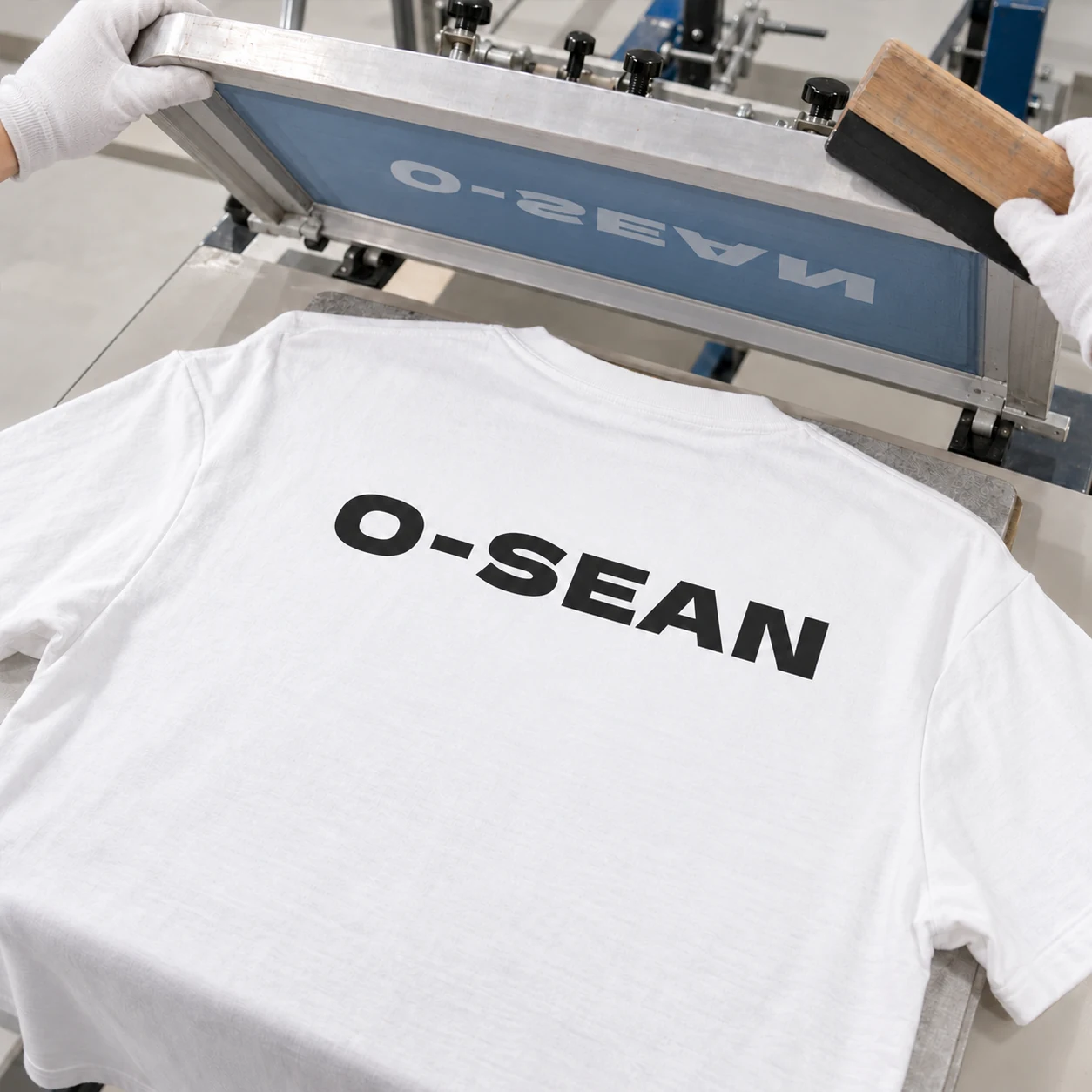 Screen printing process: a lifted screen and squeegee above a logo printed on the back of a white T-shirt